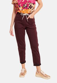 Donna che indossa pantaloni bordeaux scuri con tasche frontali e cordoncino bianco, abbinati a capelli arrotolati e scarpe da ginnastica metalliche color oro rosa.