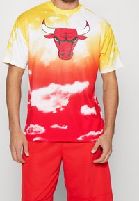 Koszulka z gradientem przechodzącym od żółtego do czerwonego oraz białymi wzorami chmur. Na środku grafika czerwonej głowy byka z białymi rogami.