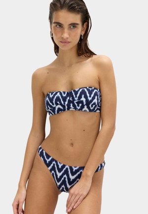 Kobieta ubrana w granatowo-biały bikini bez ramiączek z wzorem w zygzaki oraz dopasowane majtki bikini na jednolitym tle.