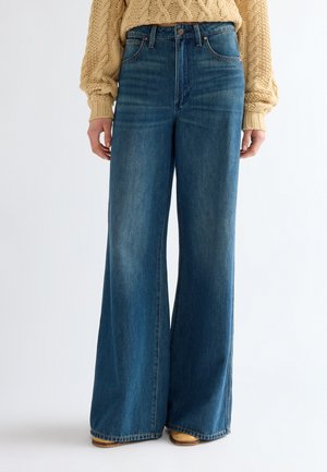 Jeans blu a gamba larga indossati con scarpe color cuoio e un maglione beige lavorato a trecce su uno sfondo chiaro uniforme.