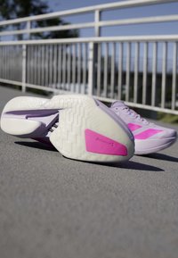Par lyse lilla og pink sneakers på tekstureret udendørs overflade nær hvid metalgelænder under klar himmel.