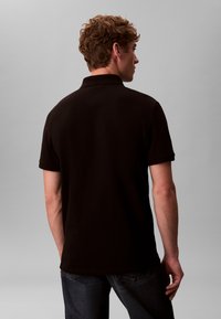 Μαύρο πόλο πουκάμισο με γιακά, κοντά μανίκια και ίσια γραμμή. Κατασκευασμένο από λείο ύφασμα με διακριτική λάμψη. Ορατή η πίσω όψη.