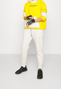 Geel en grijs hoodie met logo, gecombineerd met lichtgrijze joggers en zwarte sportschoenen. Handschoenen met een vingerloze design.