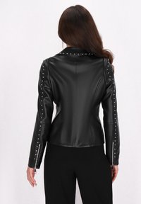 Jachetă din piele faux neagră cu un guler unghiular, accente cu ținte pe ambele mâneci și manșete cu fermoar. Textură netedă, design ajustat.
