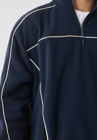 Pólder de forro polar de color navy con un cuello alto, que presenta un ribete blanco en el frente y las mangas. Cierre del cuello con cremallera.
