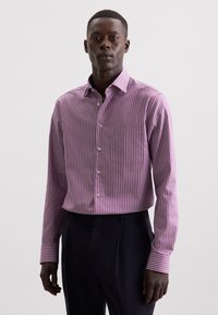 Ein Mann trägt ein lila und weiß gestreiftes Hemd mit schwarzer Hose und steht vor einem hellen, einfarbigen Hintergrund.