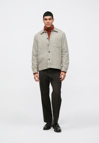 Muž v hnedom roláku, károvanom sako s gombíkmi, tmavých nohaviciach a čiernych topánkach stojí na pozadí s jednoduchou bielou farbou.