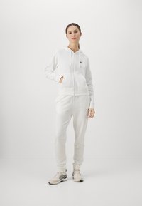 Bijela zip-up dukserica s bočnim džepovima i rebrastim manžetama, u paru s uskim trenirkama i sportskim cipelama neutralnih tonova.
