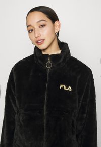 Giacca nera in pelliccia sintetica con zip, colletto alto, caratterizzata da un logo FILA dorato e un cursore della zip circolare. Texture liscia e vestibilità comoda.