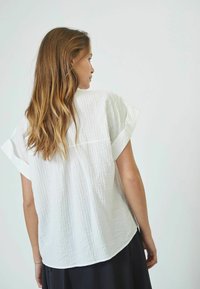 Blusa bianca a maniche corte con una texture stropicciata, vestibilità rilassata e scollo rotondo, abbinata a una gonna scura.
