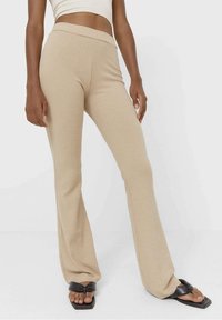 Beige geribbelde flared broek met een hoge tailleband en zachte textuur, gecombineerd met zwarte sandalen met een strikdetail.