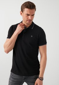 Černé polo triko s úzkým střihem, krátkými rukávy, šedými akcenty na límci a malým logem na hrudi. Texturovaná látka.