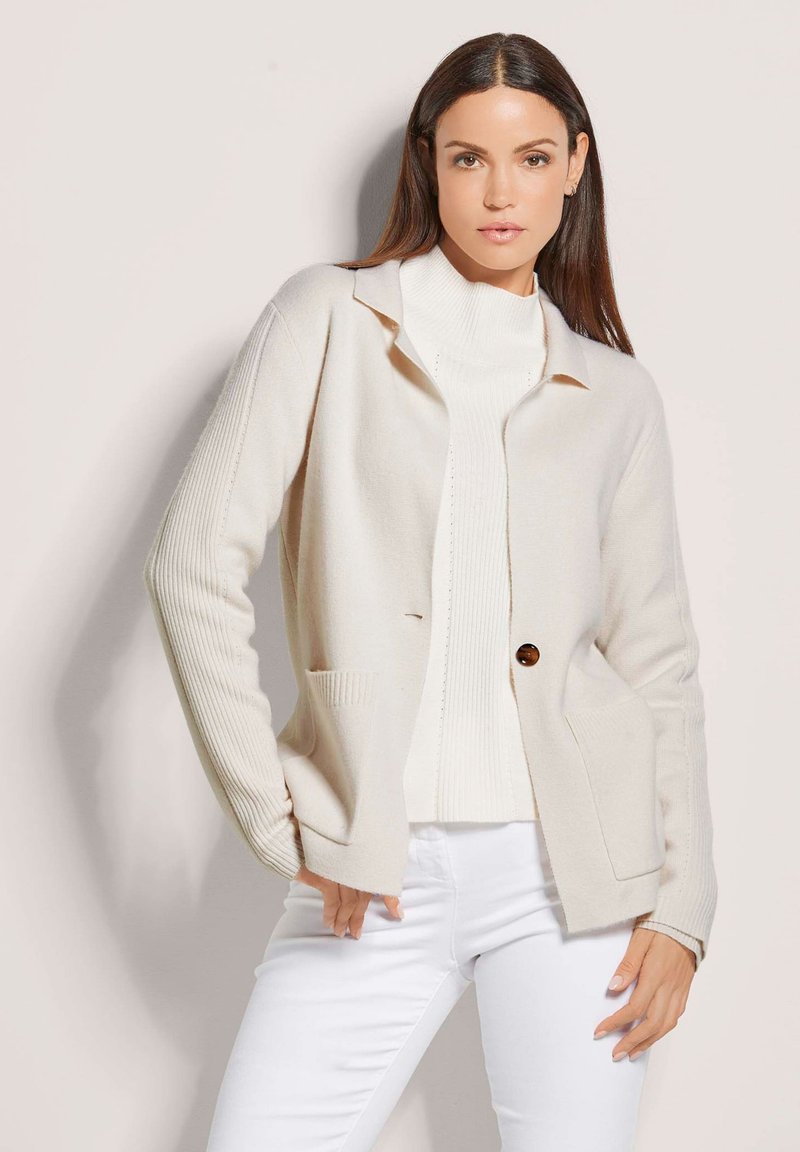 Cremefarbener Cardigan mit kragenlosem Ausschnitt, zentralen Knopfverschluss und gerippten Ärmeln, getragen über einem weißen Rollkragenpullover und weißen Hosen.