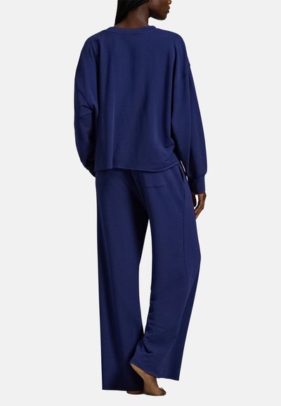 Άτομο που στέκεται ξυπόλητο, φορώντας άνετο σκούρο μπλε μπλουζάκι με μακριά μανίκια και παντελόνι με φαρδιά μπατζάκια, από πίσω, με απλό φόντο.