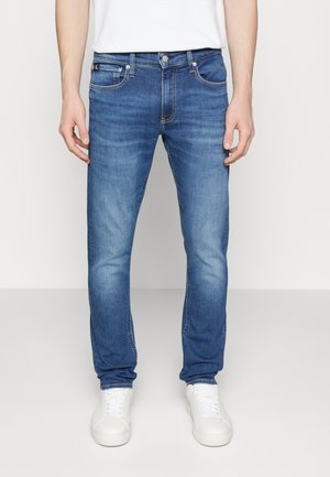 Mann trägt Slim-Fit-blaue Jeans mit leichtem Verblassen, weiße Sneaker und ein weißes T-Shirt, steht vor einem einfarbigen hellen Hintergrund.