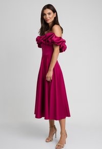 Fuksjowa sukienka na jedno ramię z falbaniastymi rękawami, dopasowanym gorsetem i rozkloszowanym dołem. Gładki materiał o lekko błyszczącej powierzchni. Szpilki.