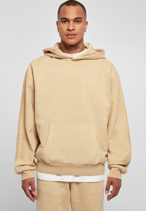 Felpa oversize beige realizzata in tessuto morbido. Presenta una tasca frontale, un cappuccio con cordino e polsini a coste. Abbinata a pantaloni della tuta beige coordinati.