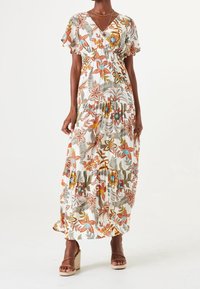Frau in einem weißen Maxikleid mit orangefarbenen, blauen und grünen Blumenmustern und rüschensäumigen kurzen Ärmeln, kombiniert mit beigen Keilsandalen.