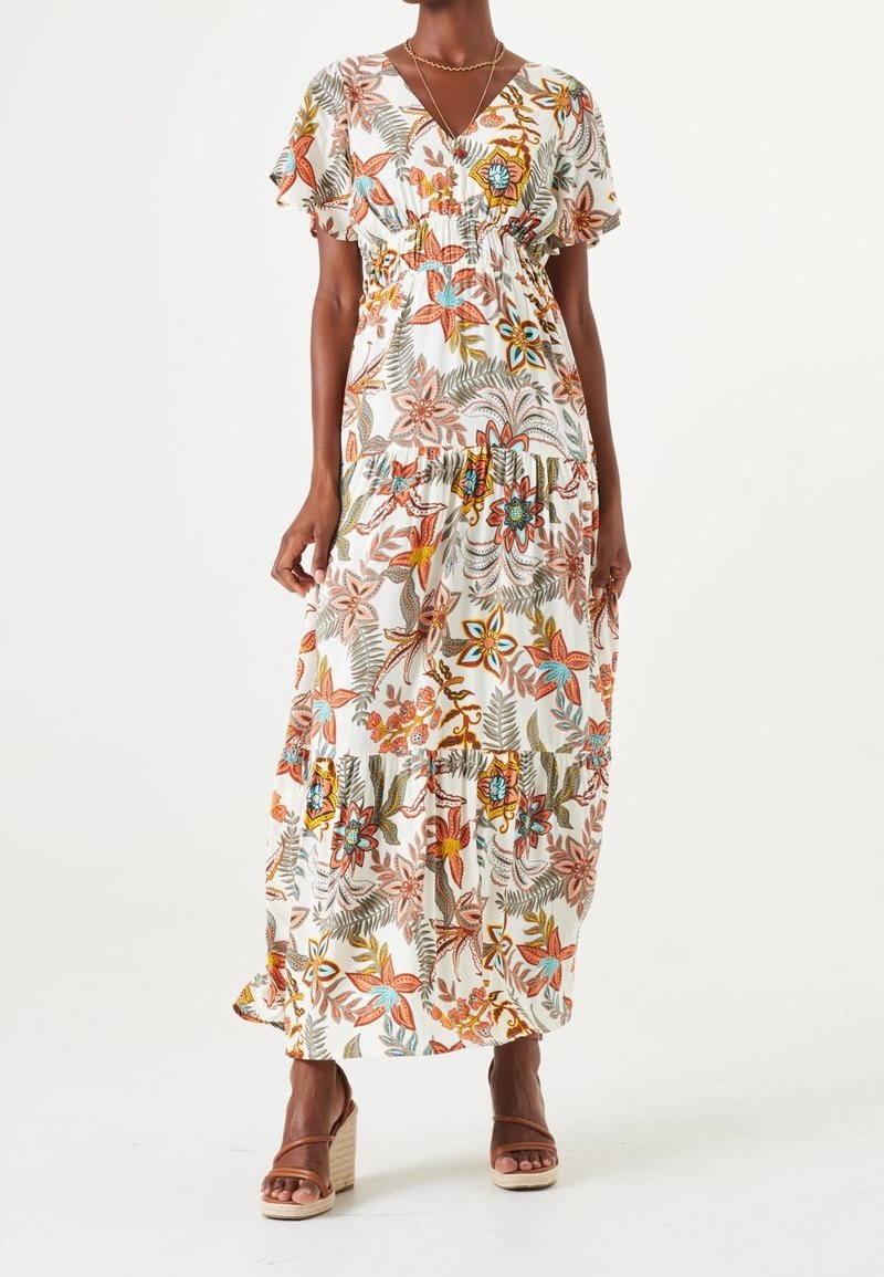 Frau in einem weißen Maxikleid mit orangefarbenen, blauen und grünen Blumenmustern und rüschensäumigen kurzen Ärmeln, kombiniert mit beigen Keilsandalen.