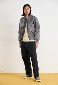 Veste bomber grise avec une fermeture éclair à l'avant, des poignets côtelés et une poche sur le bras ; portée avec une chemise beige, un pantalon sombre et des baskets noires.