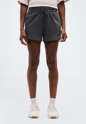 Osoba ubrana w czarne szorty z sznurkiem, białe skarpetki i białe buty sportowe, stojąca na tle jednolitego szarego tła.