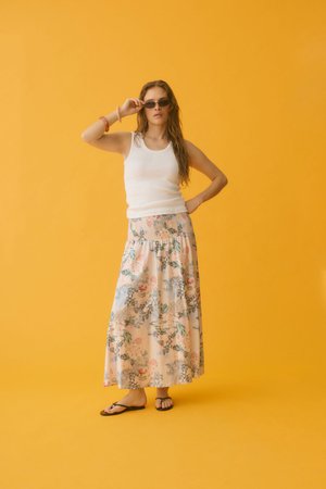 Femme aux longs cheveux portant des lunettes de soleil, un débardeur blanc, une jupe longue fleurie et des sandales, debout devant un fond jaune uni.
