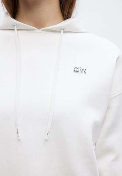 Sweat à capuche blanc avec cordons de serrage à l'avant portant l'inscription "Lacoste" et petit logo crocodile brodé sur la poitrine gauche, porté par une personne.