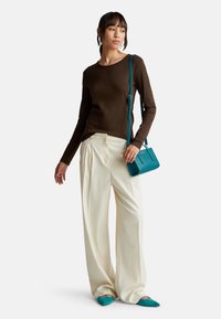 Vrouw in een bruine langärmmige top, crèmekleurige wijde broek, teal puntige ballerina's en een teal schoudertas, staande tegen een witte achtergrond.
