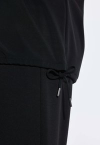 Indumento nero con cappuccio caratterizzato da un cordoncino stringente in vita con punte metalliche argentate, realizzato in tessuto leggero e liscio.