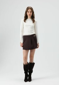 Bruine rok met plooien en gouden kettinkdetail. Wit top met lange mouwen en knoopdetails. Zwarte kniehoge laarzen. Effen grijze achtergrond.