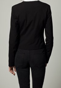 Černý přiléhavý blazer s kulatým výstřihem a upravenými švy, doplněný černými džínami s dvěma zadními kapsami a hladkou texturou.
