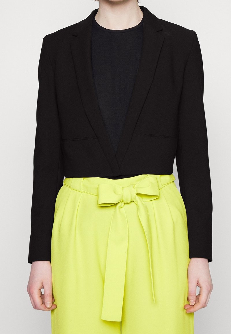 Czarna krótka marynarka z otwartym kołnierzem, w połączeniu z wysokim stanem jaskrawożółtymi spodniami z ozdobą w formie wiązanej kokardy.