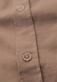 Tissu d'une chemise marron à boutonnière avec une fine texture. Dotée d'un bouton blanc à quatre trous et de coutures contrastées le long de la patte de boutonnage.