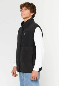Chaleco de forro polar negro con cuello alto, bolsillo con cremallera en el pecho y bolsillos laterales. Llevado sobre una camiseta de manga larga blanca, con un corte casual.