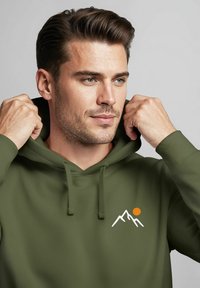 Olive green hoodie made of soft fabric, featuring a white mountain and orange sun graphic on the chest. Adjustable drawstring hood.