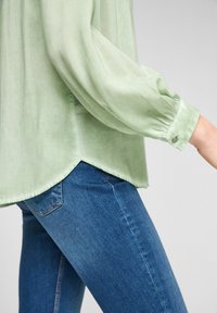 Blusa leggera verde chiaro, trasparente, con maniche lunghe a sbuffo, con orlo arrotondato e polsini con bottoni, abbinata a jeans in denim blu scuro.
