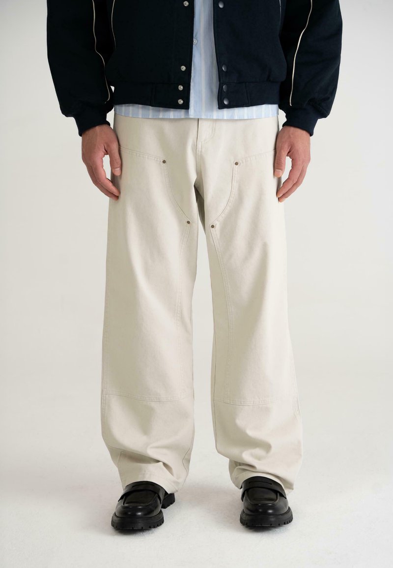 Pantalons larges beige avec une texture lisse, dotés de deux poches avant et de coutures visibles, associés à une veste noire et des chaussures sombres.
