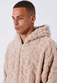 Fleecia con cappuccio beige, caratterizzata da un motivo ondulato. Presenta una zip frontale e un materiale morbido e peloso per calore e comfort.
