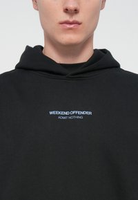 Sort hættebluse lavet af blødt materiale. Har en hætte med snører og et centralt tryk på brystet i lyseblå: "WEEKEND OFFENDER ADMIT NOTHING."