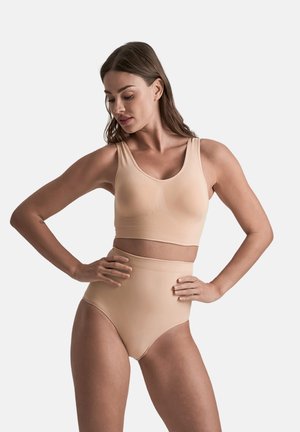Donna con lunghi capelli castani che indossa biancheria intima beige a vita alta senza cuciture e un reggiseno sportivo abbinato, in posa con le mani sui fianchi su uno sfondo bianco.