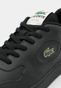 Baskets en cuir noir à forme arrondie, panneau latéral texturé, lacets noirs et logo vert brodé sur le côté extérieur.