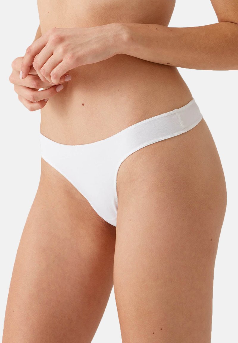 Witte stringonderbroek van zacht materiaal met een gladde textuur. Heeft een dunne elastische tailleband, minimale bedekking en een naadloos ontwerp.