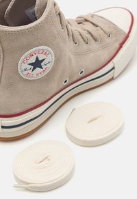 Zapatilla alta de ante beige con punta de goma blanca, suela a rayas rojas y azules, y el icónico logo de estrella. Incluye dos cordones blancos.