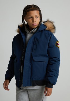Junge Person mit geflochtenem Haar, die eine blaue Winterjacke mit Fellkapuze und einem Aufnäher am Ärmel trägt und zur Seite schaut.