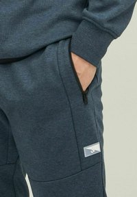 Mörkblå joggingbyxor i mjukt tyg, med en ficka med dragkedja och en liten logotyp med vit bakgrund på sidan.