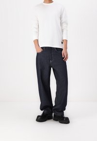 Dunkelblaue, locker sitzende Jeans aus Denim mit sichtbaren Nähten, kombiniert mit einem weißen Langarmshirt und schwarzen Schuhen mit klobiger Sohle.