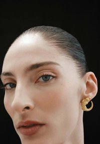 Pendiente de aro dorado con diseño texturizado de cocodrilo, de tipo clip. El retrato en primer plano muestra una piel suave y cabello liso contra un fondo oscuro.