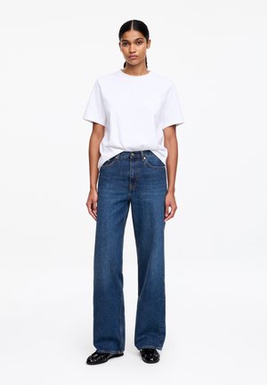 Jauna moteris su tamsiais plaukais, surištais žemu kuoduku, dėvi paprastą baltą trumpomis rankovėmis marškinėlį, laisvas mėlynas džinsines kelnės ir juodus batus, stovinti priešais baltą foną.