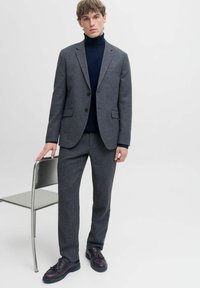Siva vunena blejzer s izrezanim reverima, turtleneck od mrežaste tkanine, usklađene sive hlače i tamne kožne cipele. Stoji pored metalne stolice.