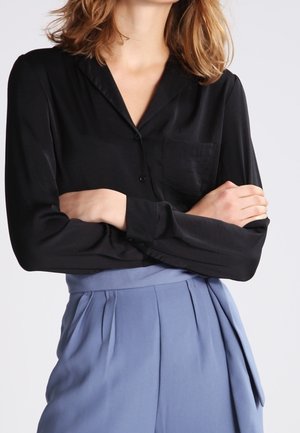 Kobieta z założonymi rękami, ubrana w czarną bluzkę z długim rękawem i plisowane, niebieskie spodnie z wysokim stanem na jednolitym tle.